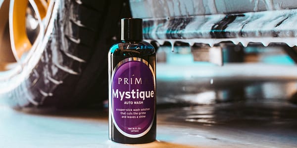 A bottle of Prima Mystique auto wash sits in front of a car dripping with soapy water