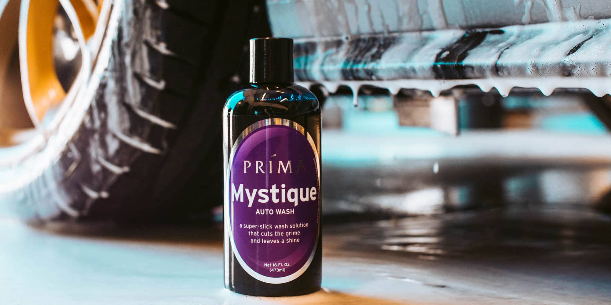 A bottle of Prima Mystique auto wash sits in front of a car dripping with soapy water